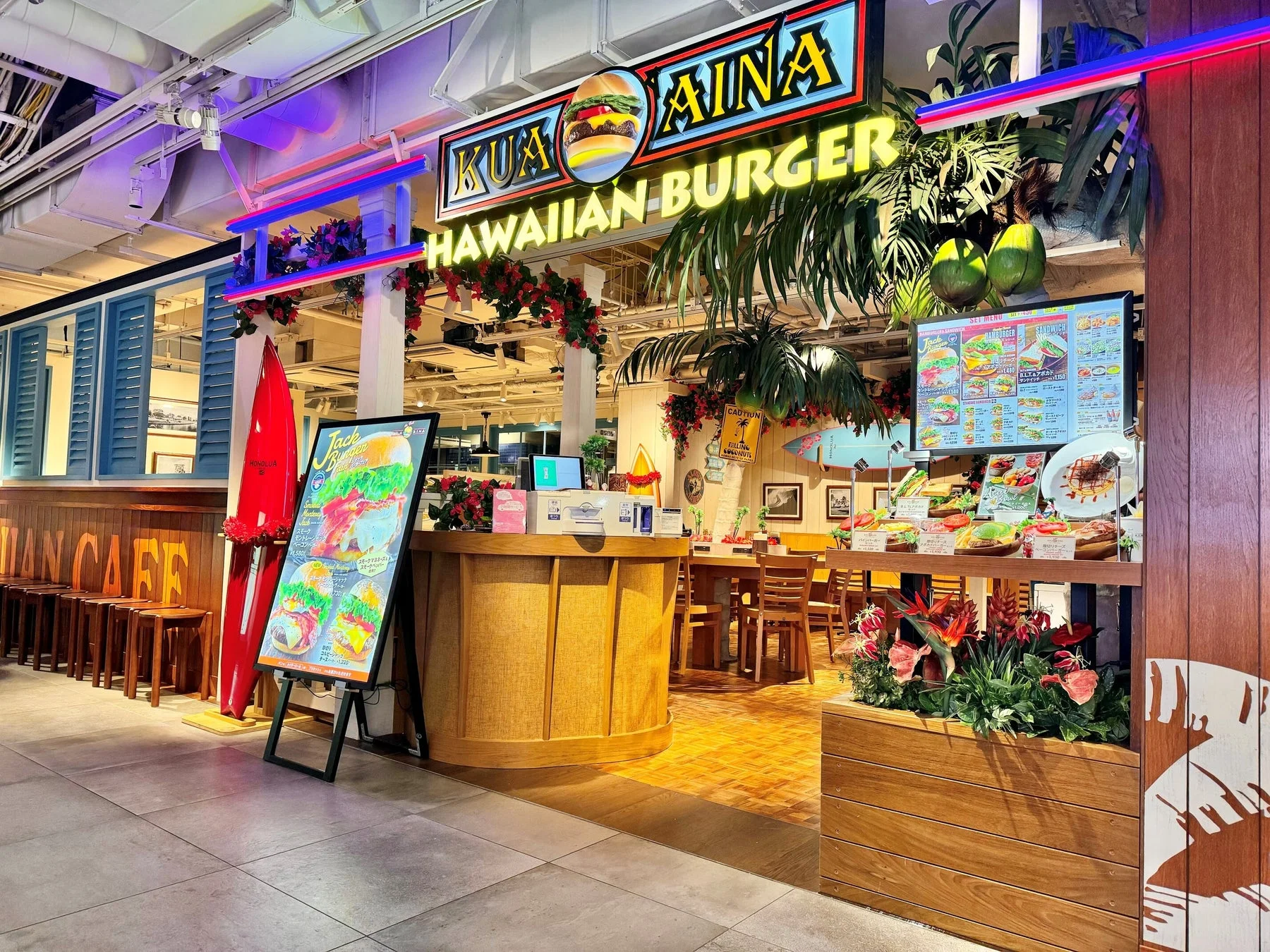 KUA`AINAというハワイアンバーガー店の外観です。店内はヤシの木やサーフボード、ハワイアンな装飾で飾られ、メニューディスプレイにはハンバーガーやサンドイッチが並んでいます。ネオンサインが特徴的な、南国リゾートのような雰囲気のお店です。