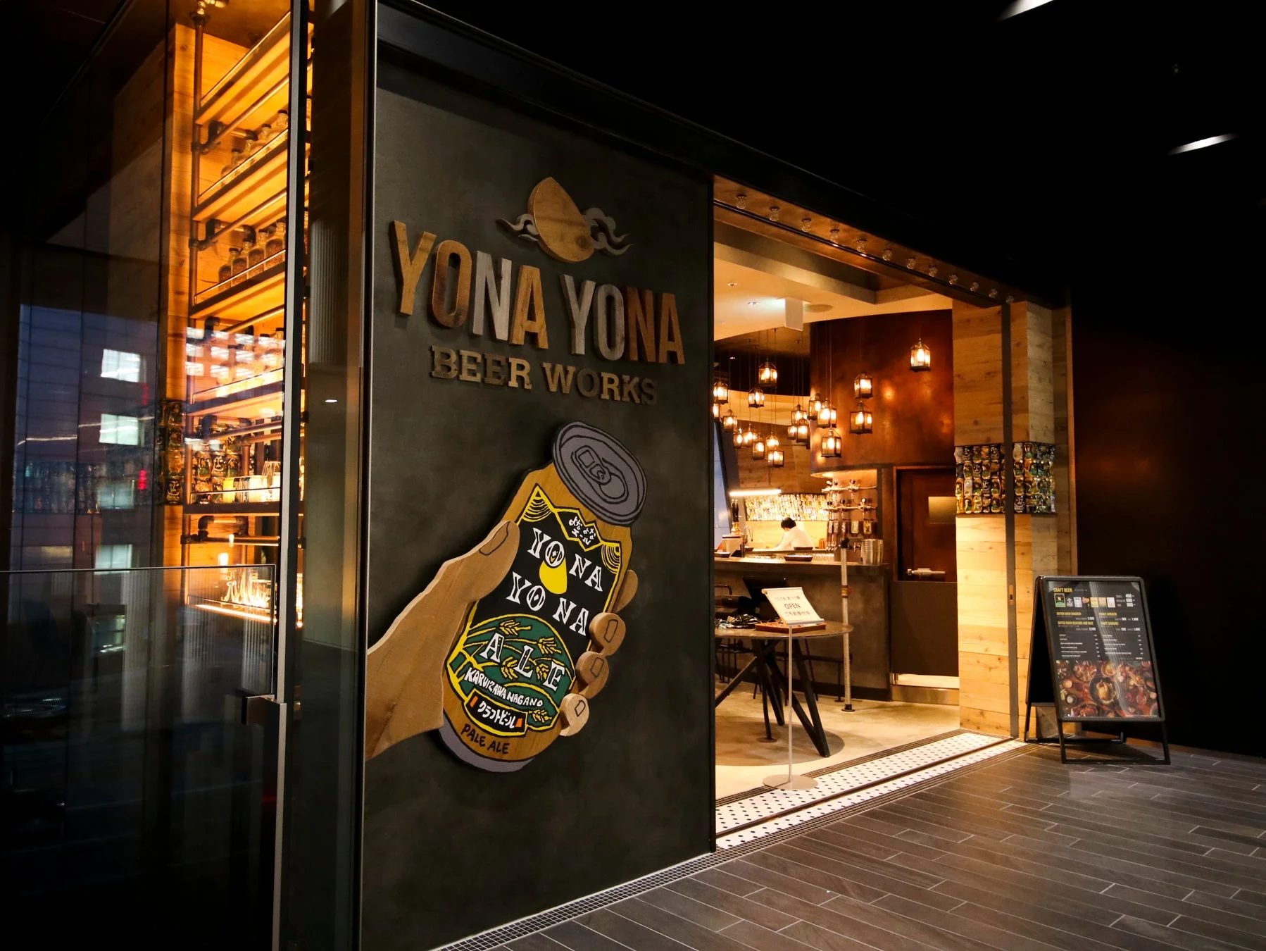 「YONA YONA BEER WORKS」の入り口を捉えた画像です。大きなビール缶のイラストが特徴的で、店内には温かい光のランタンが多数吊るされ、活気ある雰囲気が伝わってきます。