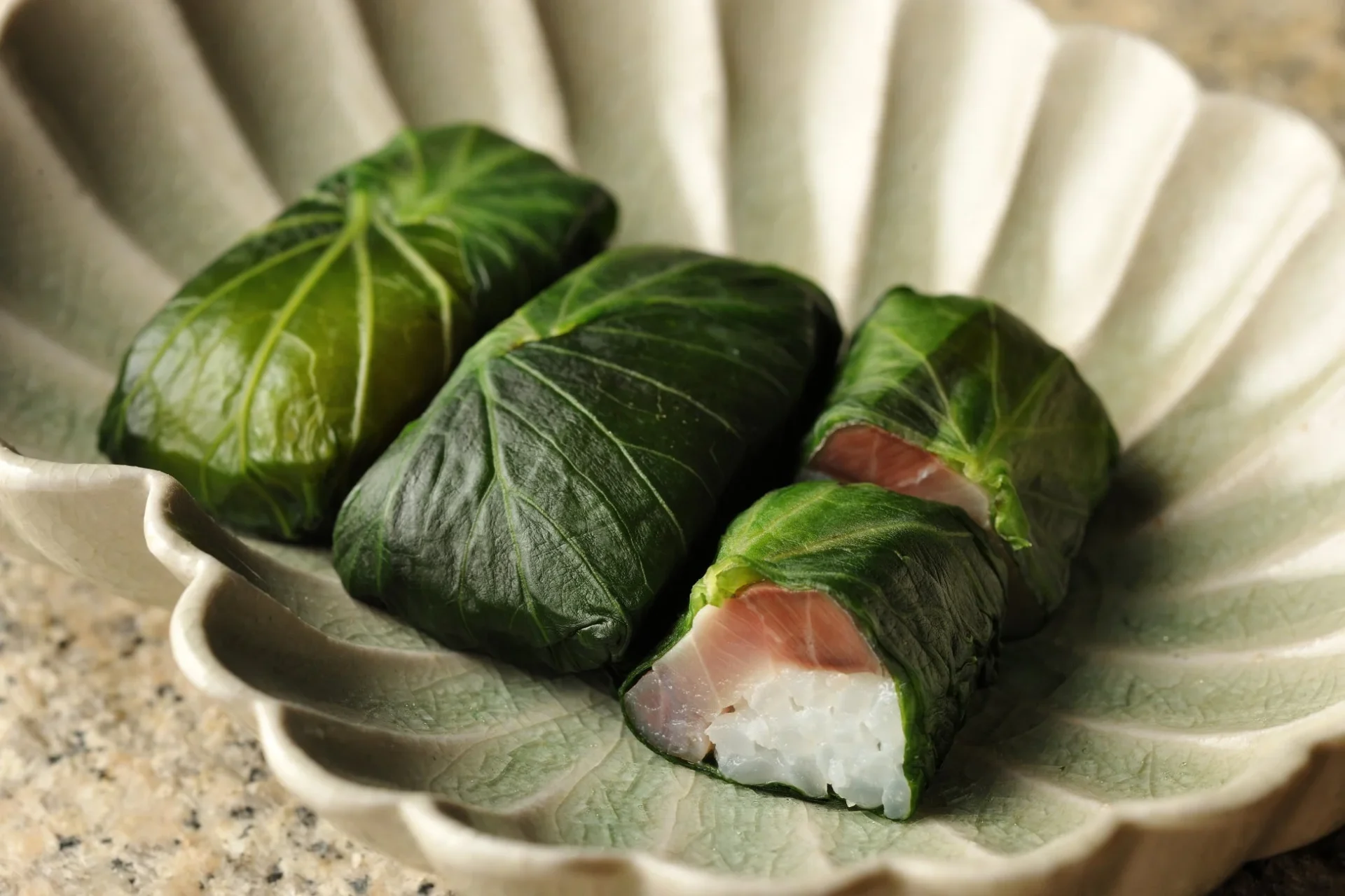 緑の葉で包まれた数切れの料理が、波打つデザインの白い陶器の皿に盛られています。一部の包みからは、白いご飯とピンク色の魚の切り身が見えており、彩り豊かで美味しそうな一品です。