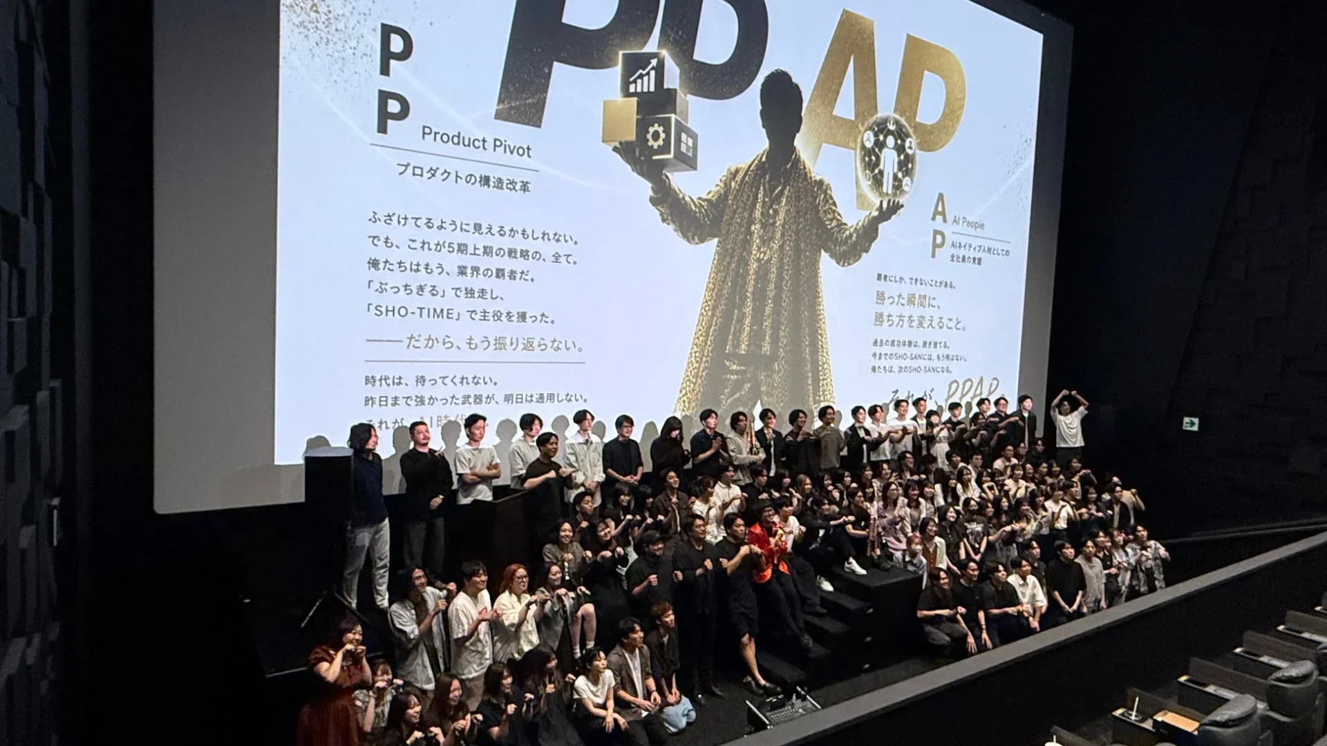 大勢の社員が会場で、企業戦略やAIネイティブ人材への変革をテーマにしたプレゼンテーションが映し出されたスクリーンを背景に集合写真を撮っている。重要なビジネスイベントの様子。