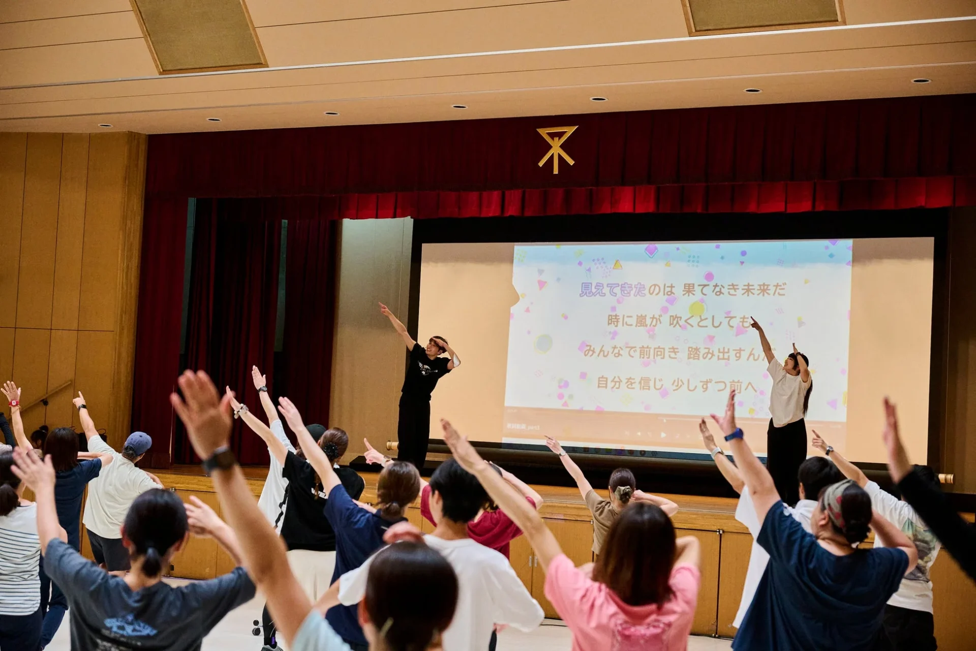 屋内で多くの人々がインストラクターの指導のもと、スクリーンに表示された歌詞に合わせて腕を上げ、楽しそうに運動している様子。