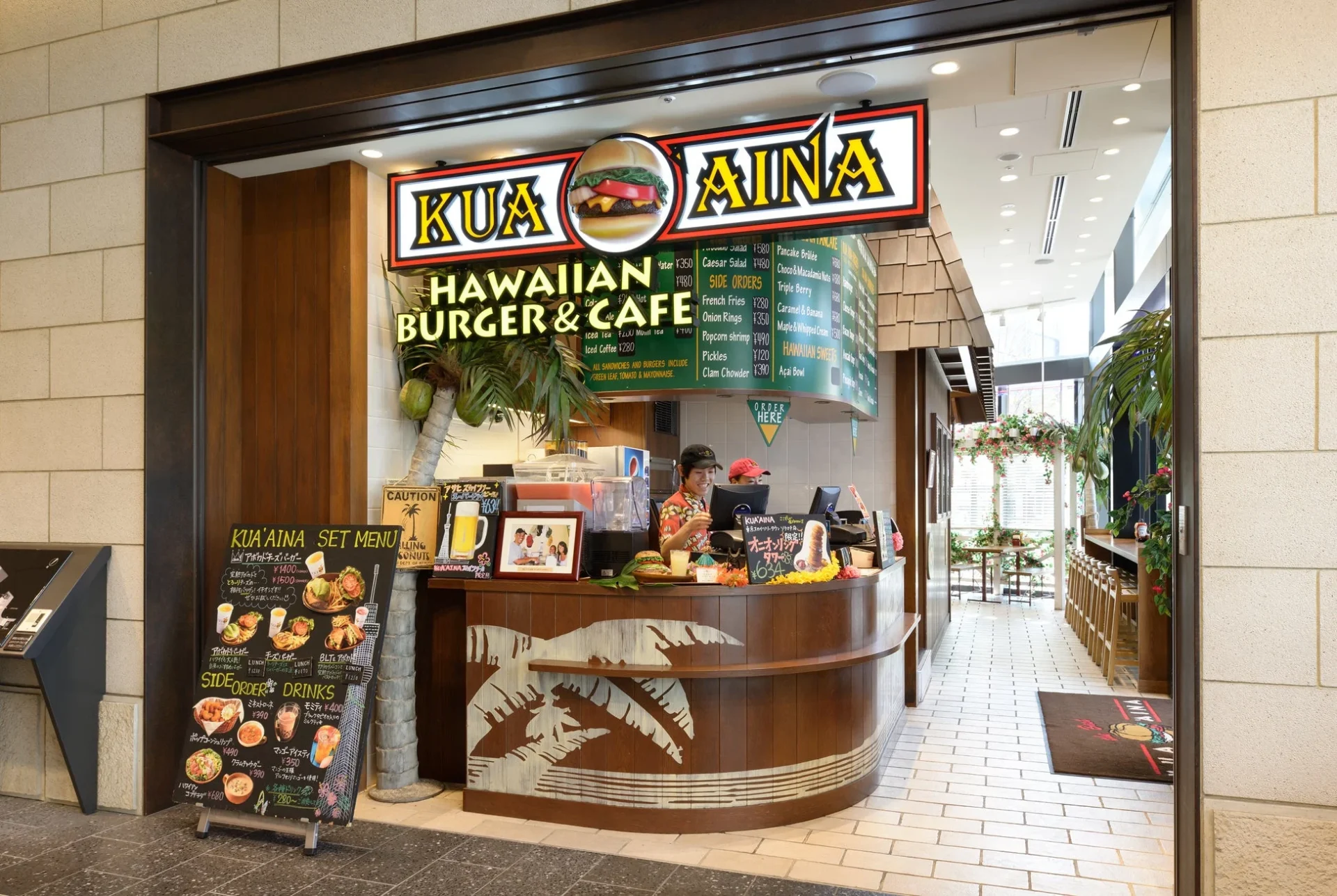 ハワイアンバーガーとカフェ「KUA AINA」の店舗外観とカウンター。店内は南国風の装飾が施され、ハンバーガーやドリンク、サイドメニューが並ぶ。スタッフが接客しており、食事を楽しめる空間が広がっている。