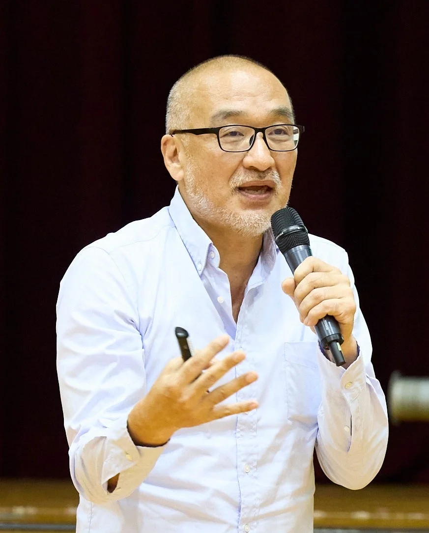 眼鏡をかけた髭のある男性が、ライトブルーのシャツを着てマイクを持ち、熱心に講演やプレゼンテーションを行っています。