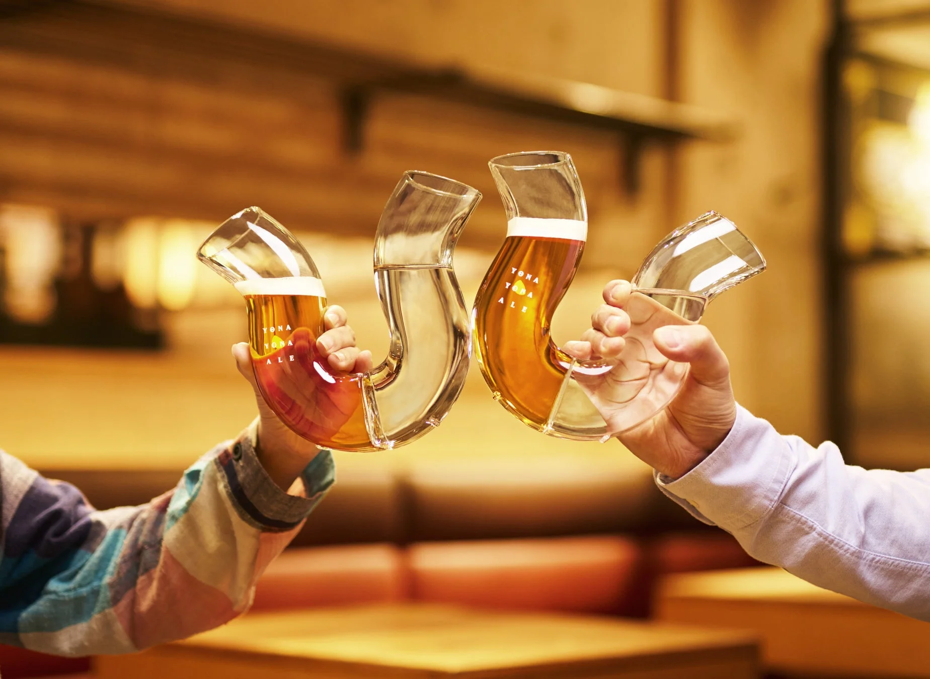 ユニークな形状のグラスでビールや水を乾杯する二人の手元を捉えた画像。ビールグラスには「YONA YONA ALE」の文字が見え、温かい雰囲気の中で楽しい交流の瞬間が表現されています。