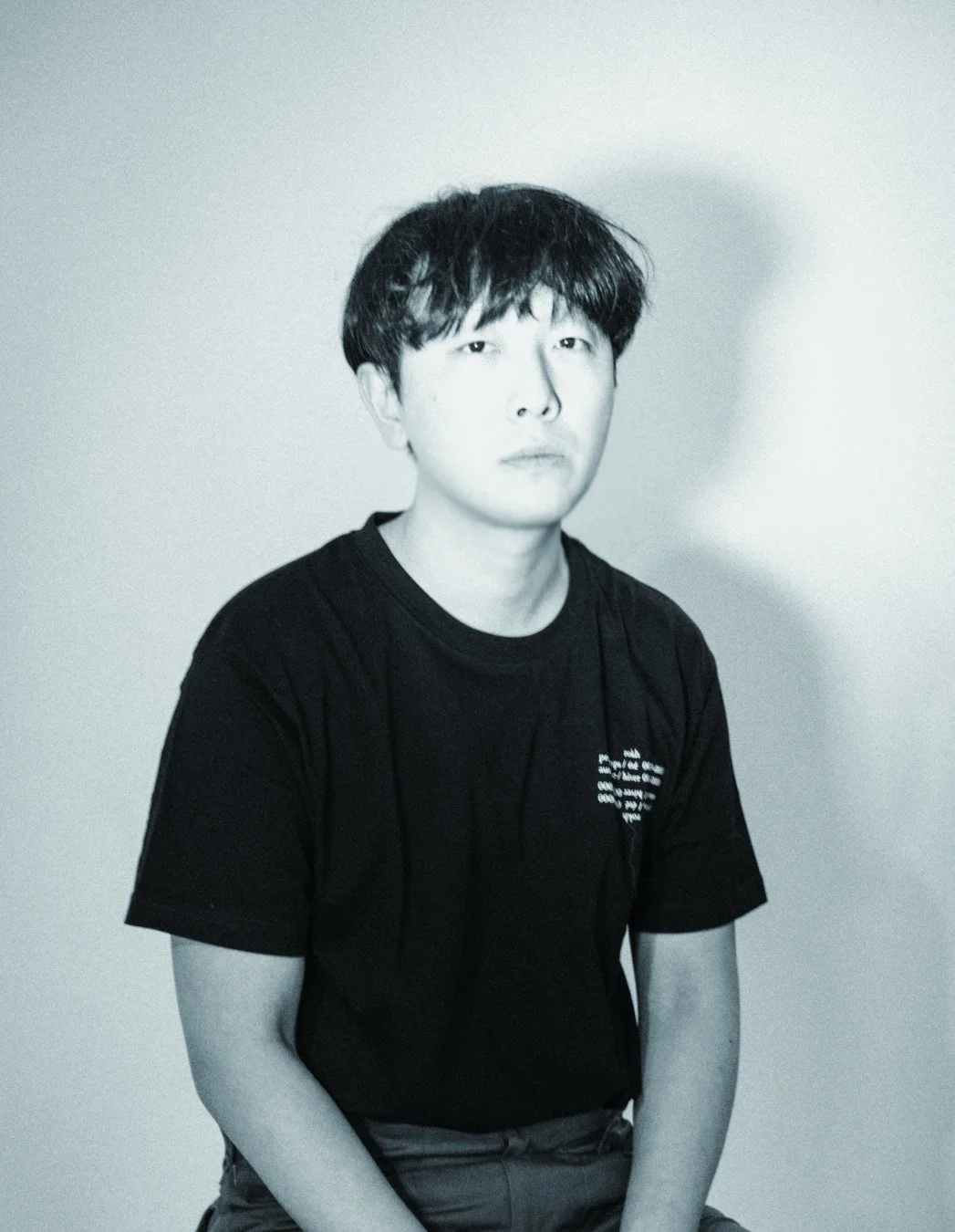 モノクロ調の画像で、黒いTシャツを着た若いアジア人男性がカメラをまっすぐ見つめて座っています。
