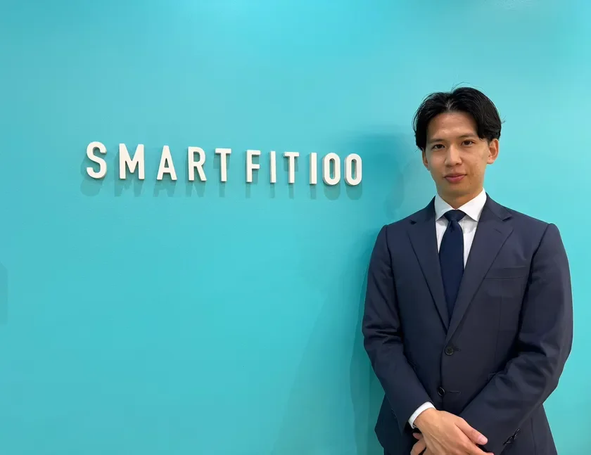 青い壁を背景に、ネイビーのスーツを着た男性が立っています。壁には「SMARTFIT100」という白い文字が書かれており、男性はプロフェッショナルな印象を与えています。
