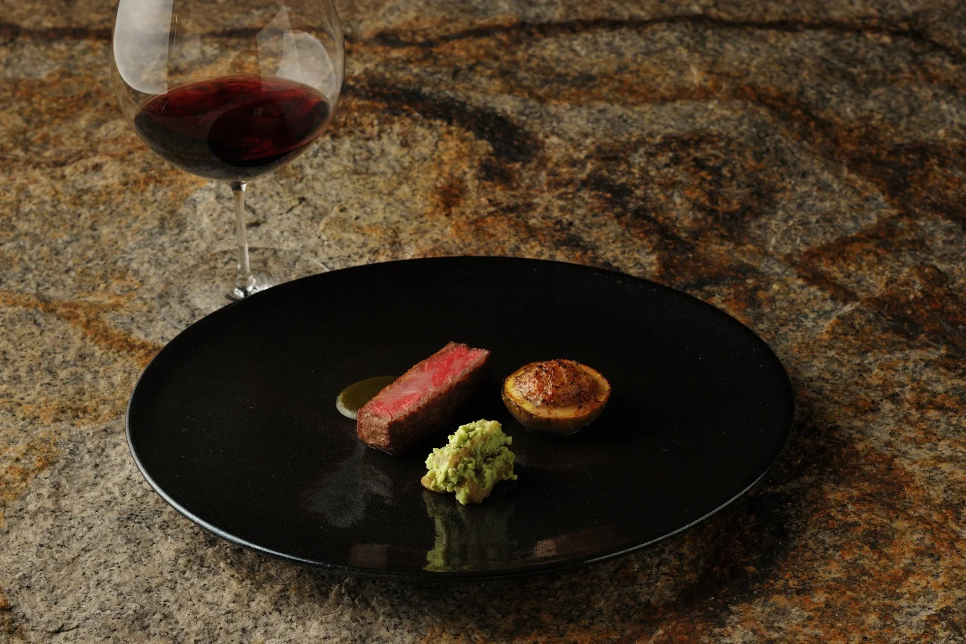 黒い皿に盛り付けられたミディアムレアのステーキと、焼きポテト、緑色のペースト。左上には赤ワインのグラスが置かれ、石のテーブルが高級感を演出しています。