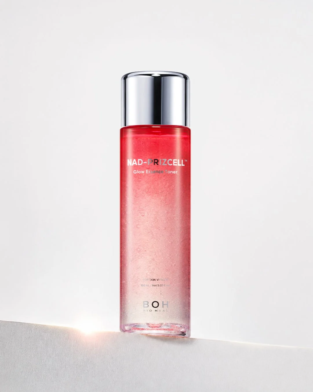 BOH BIOHEALの「NAD-PRIZCELL Glow Essence Toner」のボトルが写っています。透明感のある赤みがかったピンク色の液体が入っており、銀色のキャップが特徴です。白い台座の上に置かれ、下からの光が当たっています。