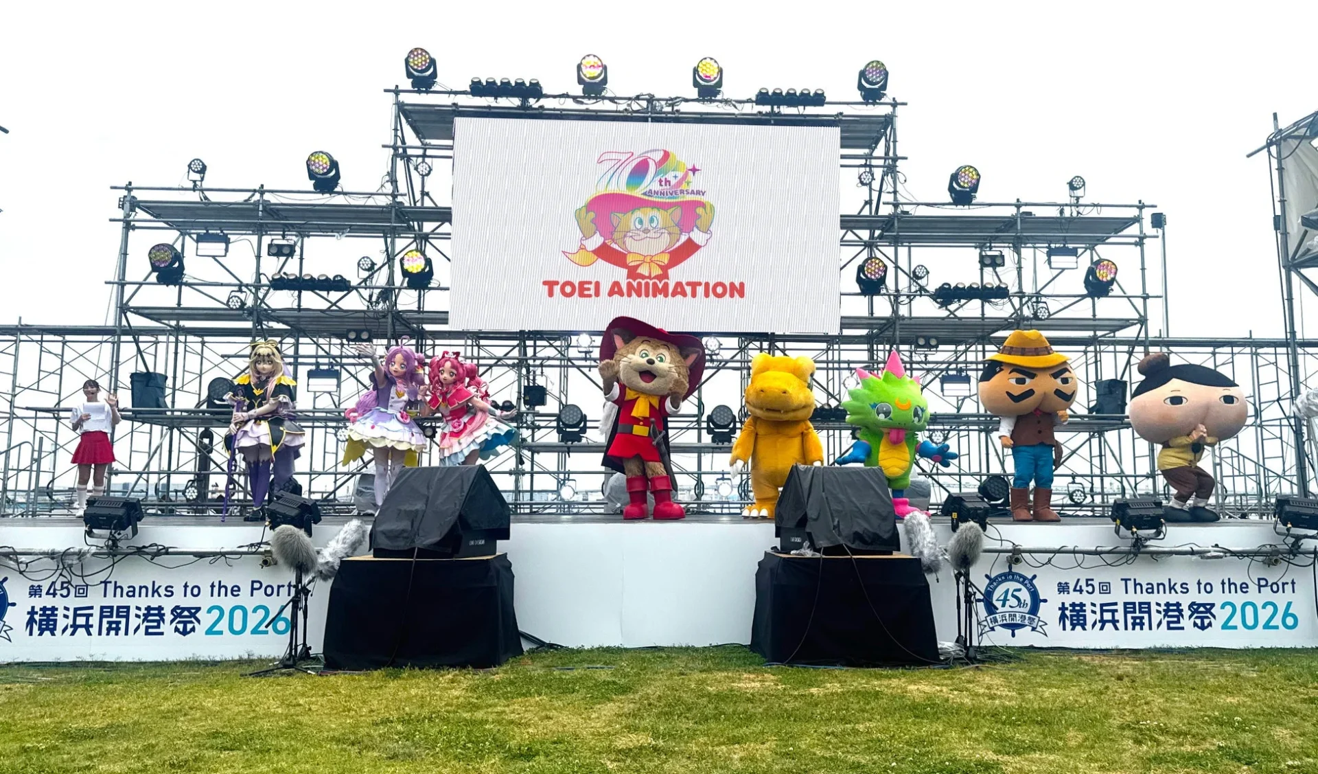 屋外ステージで東映アニメーションの70周年記念イベントが開催されており、プリキュアや長靴をはいた猫などの人気キャラクターの着ぐるみが登場している。ステージ下には「第45回 横浜開港祭 2026」と書かれたバナーが見える。