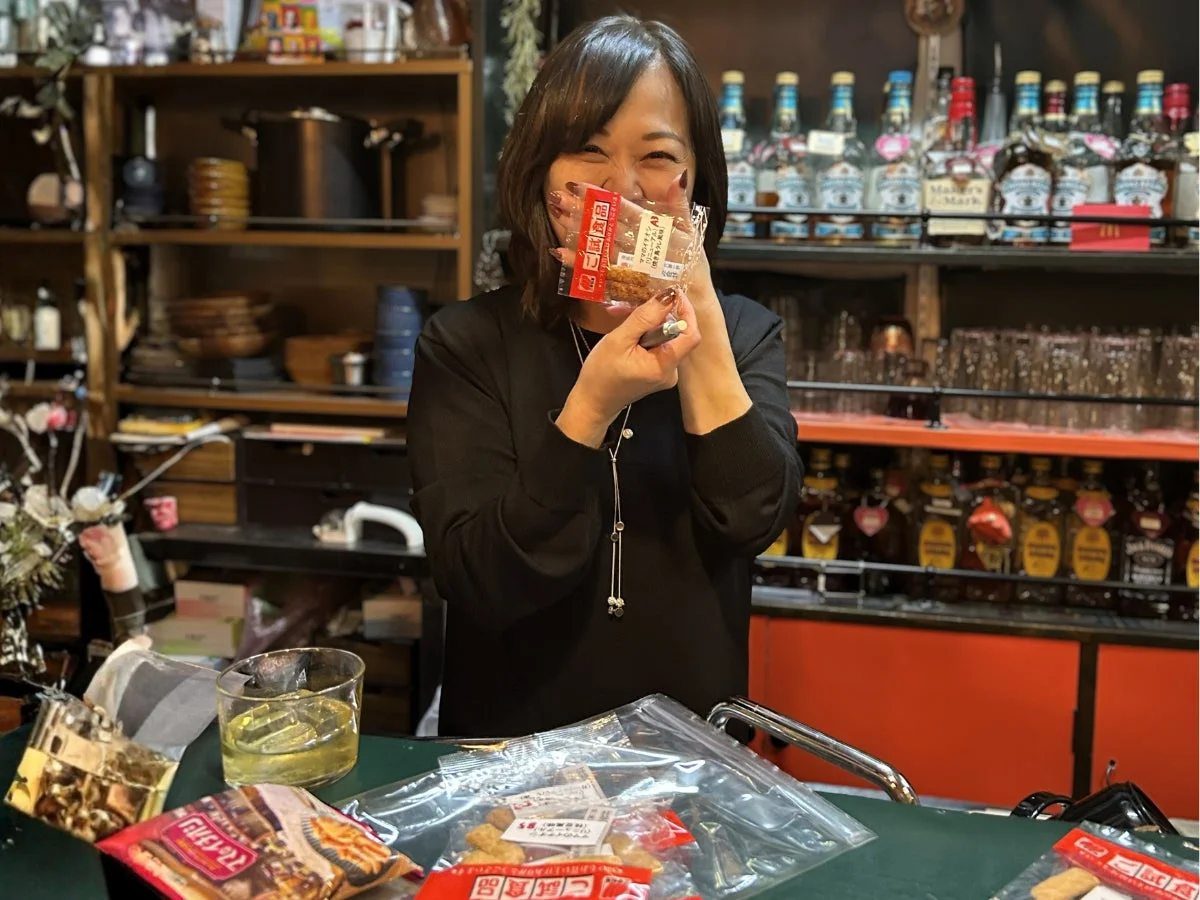 バーのような店内で、笑顔の女性がマヨネーズ風味の揚げ餅と思われるスナック菓子を手に持っている。背景には多数の酒瓶が並び、楽しそうな雰囲気が伝わってくる。