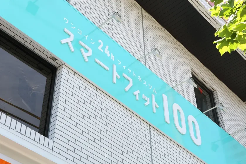 白いレンガ調の建物に設置された、ターコイズブルーの看板が写っています。看板には「ワンコイン 24h フィットネスジム スマートフィット100」と書かれており、24時間営業のフィットネスジムであることがわかります。