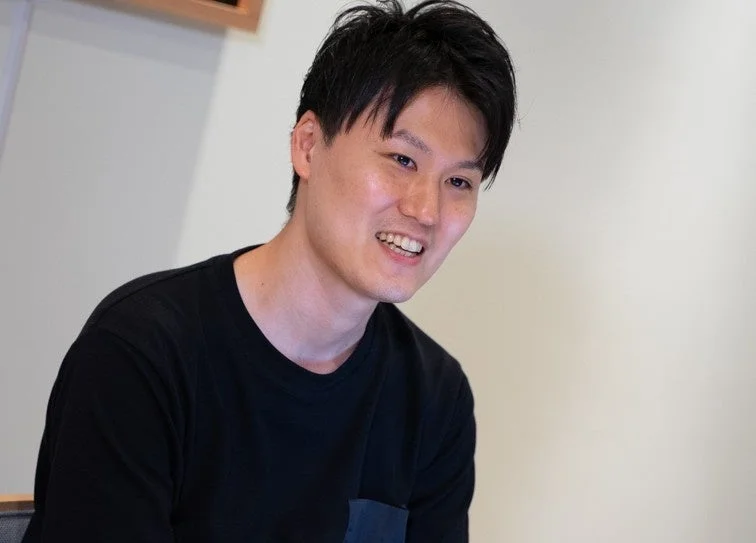 黒いTシャツを着たアジア人男性が、明るい室内で笑顔を見せているポートレート写真