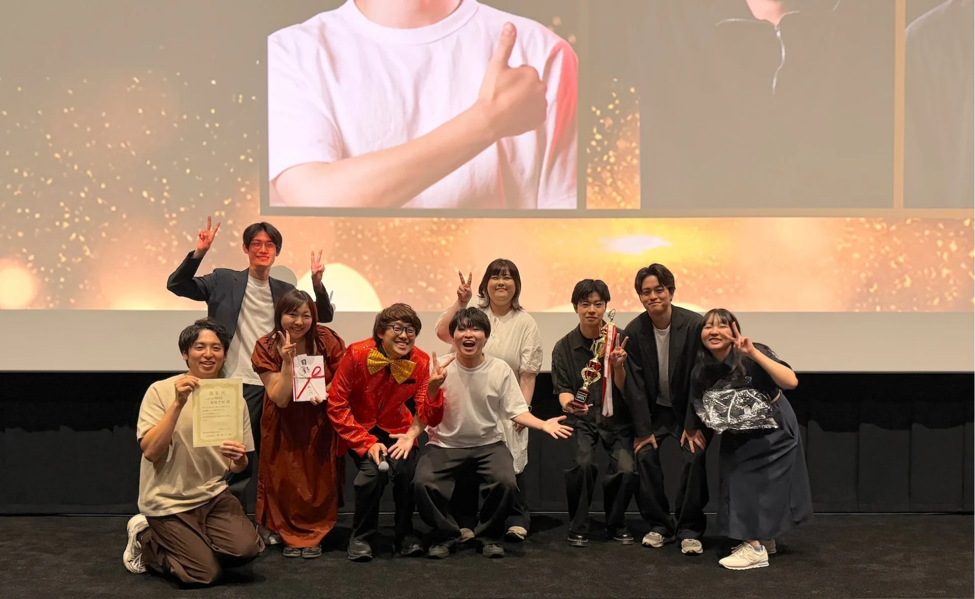 ステージで撮影された集合写真。参加者たちはトロフィーと表彰状を持ち、笑顔でピースサインをしています。何かの表彰式かイベントでの記念撮影のようで、喜びの雰囲気が伝わってきます。