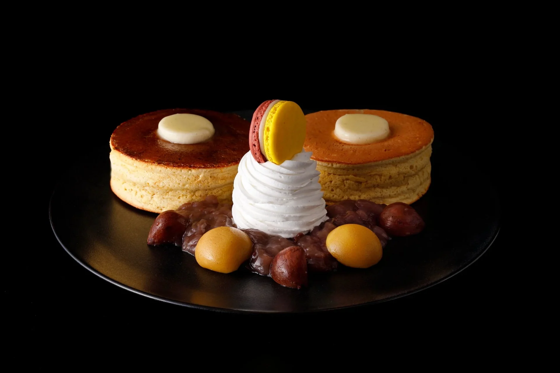 黒い背景に置かれた黒い皿に、2つのスフレパンケーキが盛り付けられています。中央にはホイップクリームとマカロン、あんこ、栗が添えられています。