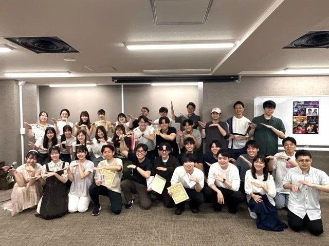 室内で多くの若者たちが笑顔で集合写真を撮っている様子です。男女混合のグループで、多くの人が特徴的な手のポーズをとっており、数名が証明書のようなものを持っています。