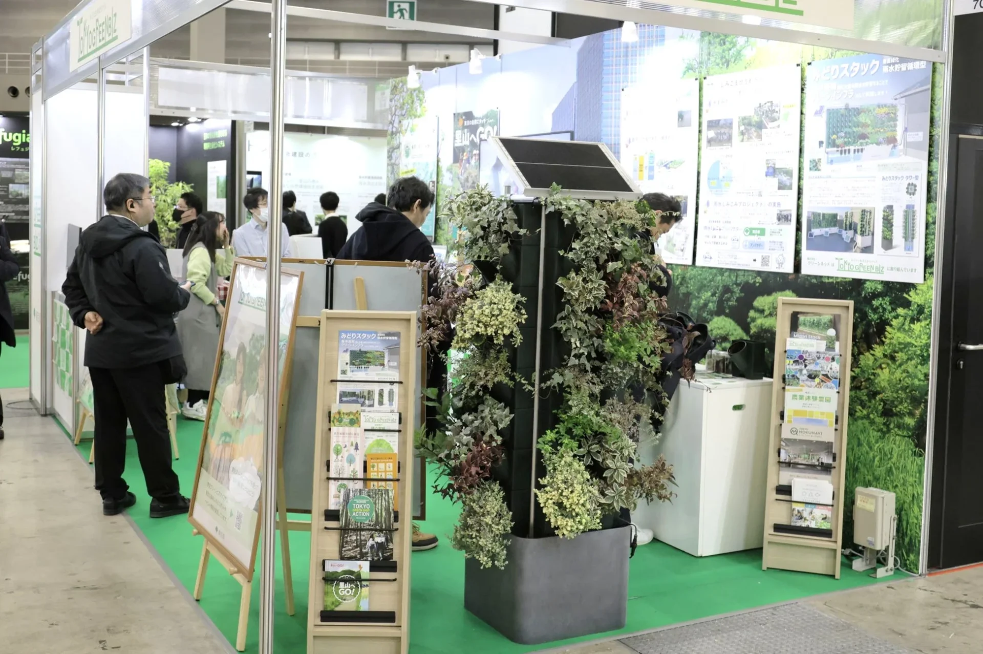 都市緑化や環境技術に関する展示会の様子。ソーラーパネル付きの大型垂直庭園が目を引くブースで、来場者が資料を見ている。みどりスタックやTokyo Green Bizといった緑のインフラソリューションが紹介されている。